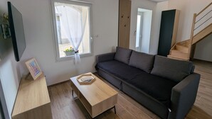 Een moderne woonkamer met een grijze bank, een houten salontafel en een raam met dunne gordijnen.