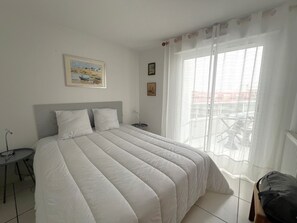 Ein Schlafzimmer mit einem großen Bett, einem kleinen Nachttisch, einem Sessel und einem Fenster mit Vorhängen.