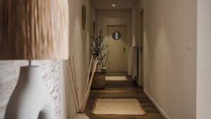Een hal met houten vloer, een witte muur met een structuur, een ronde spiegel en een plant in een pot.
