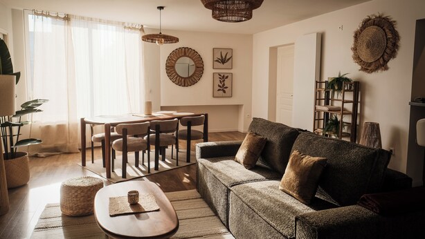 Een woonkamer met een bank, salontafel en een eethoek met stoelen. Er is een kroonluchter, een spiegel en wanddecoratie.