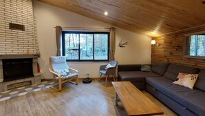 A cozy living room with a fireplace, wooden ceiling, and a large window offering a view of the outdoors.
