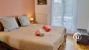 A bedroom with a bed, pillows, a nightstand with a lamp, a painting on the wall, and a window with a view of outdoor furniture. - Bright Gîte near Laval, 3 bedrooms, large grounds, ideal for families (L'Huisserie)