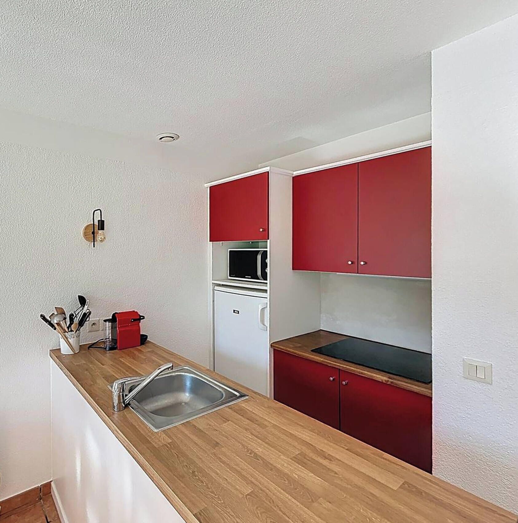 Una cocina compacta con encimera de madera, un lavamanos, armarios rojos, un microondas y un refrigerador pequeño.