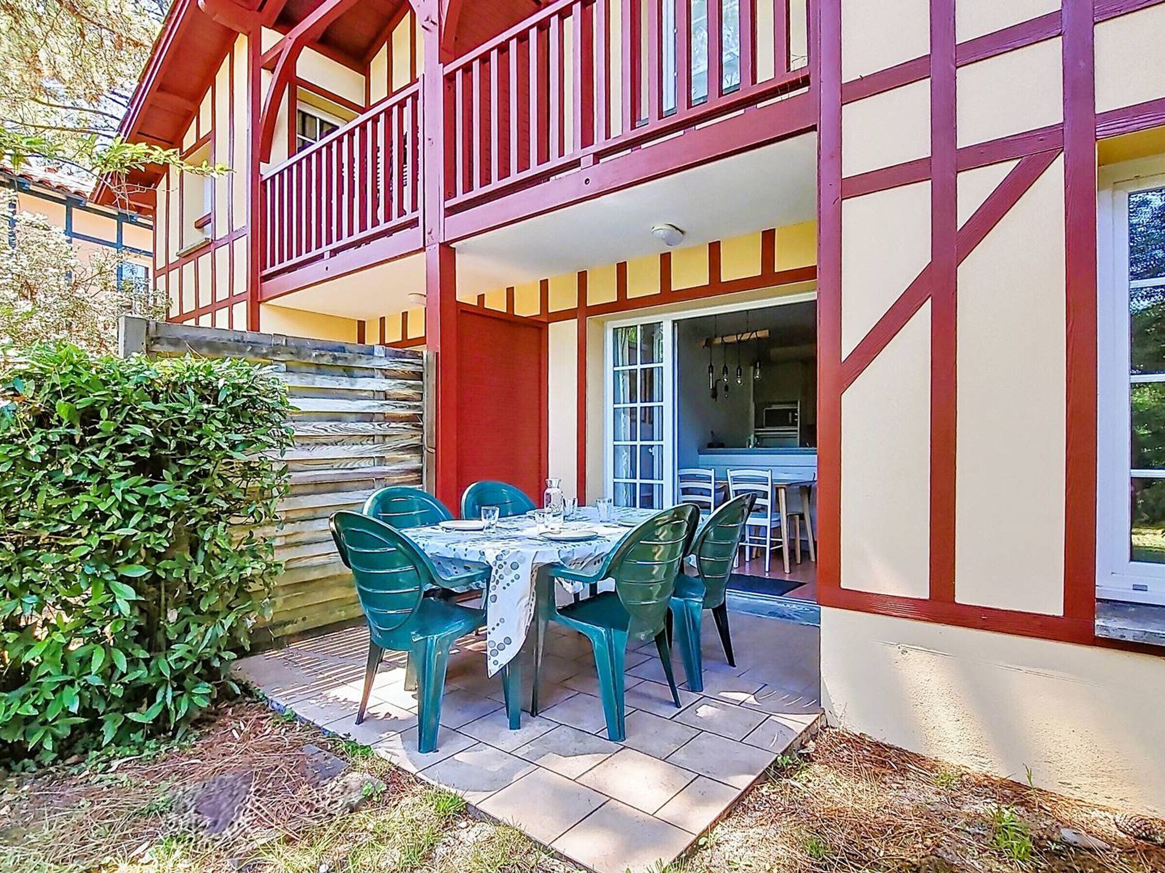 A two-story building with a balcony, a dining area with a table and chairs, and a kitchen visible through an open door.