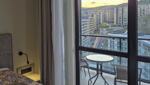 Habitación de hotel con una ventana grande, una cama, una mesita de noche y vistas a un paisaje urbano.