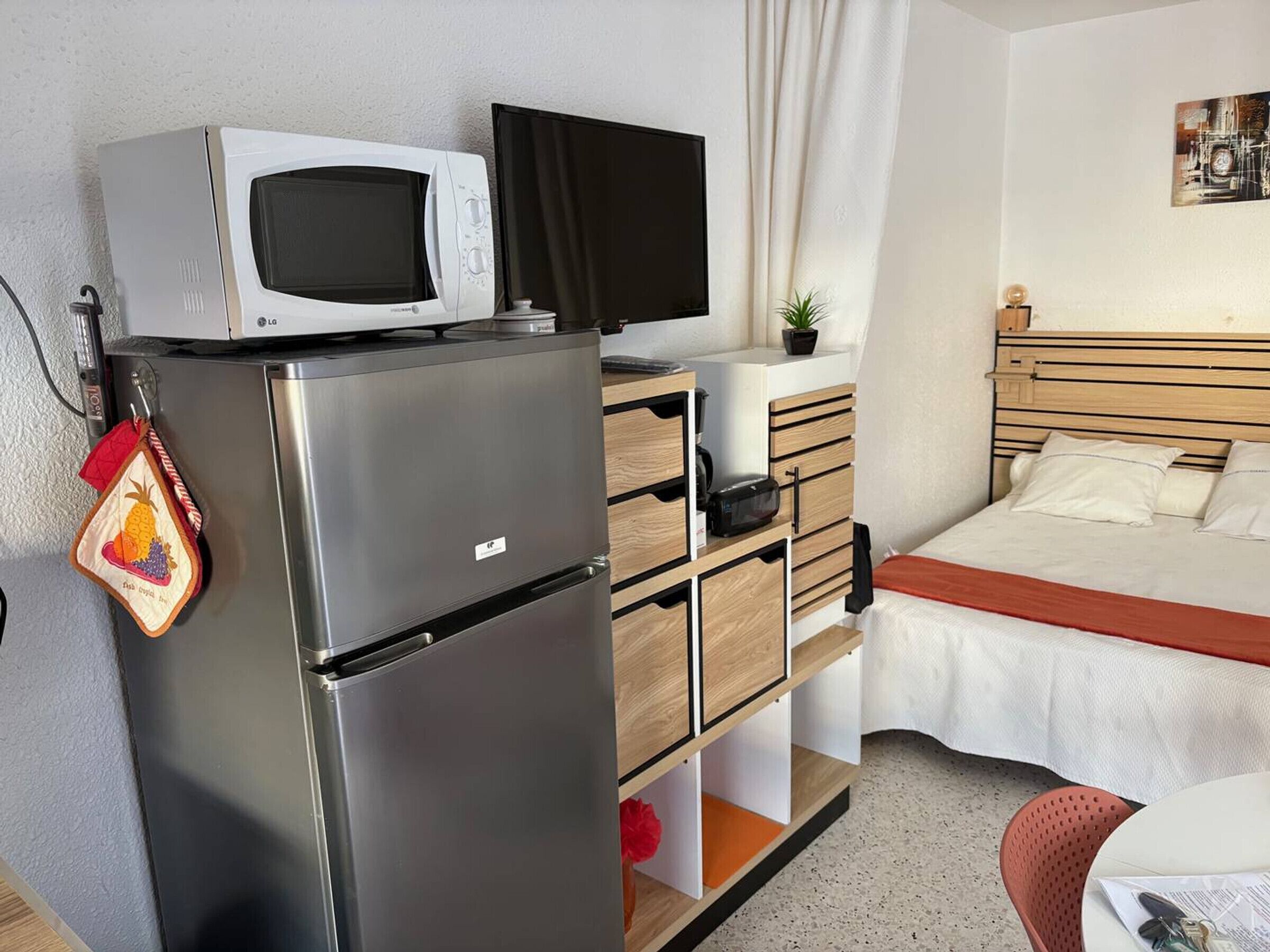 Quarto de hotel compacto com uma cama, uma escrivaninha pequena com televisão, um frigorífico e uma área de kitchenette.