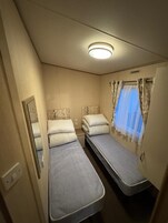 Habitación pequeña con dos camas individuales, un espejo, una ventana con cortinas e iluminación en el techo.