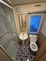 Un baño pequeño con cabina de ducha de vidrio, lavabo, inodoro y ventana con persianas.