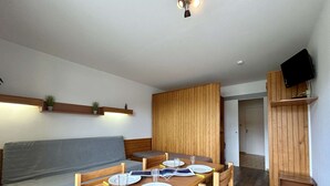 Un salon moderne avec une table à manger en bois, préparée pour quatre personnes, un canapé gris, un lambris en bois et un téléviseur fixé au mur.