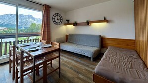 Une pièce comprenant une table à manger en bois, des chaises, un canapé, un lit, une horloge et une vue sur les montagnes, visible à travers une grande fenêtre.