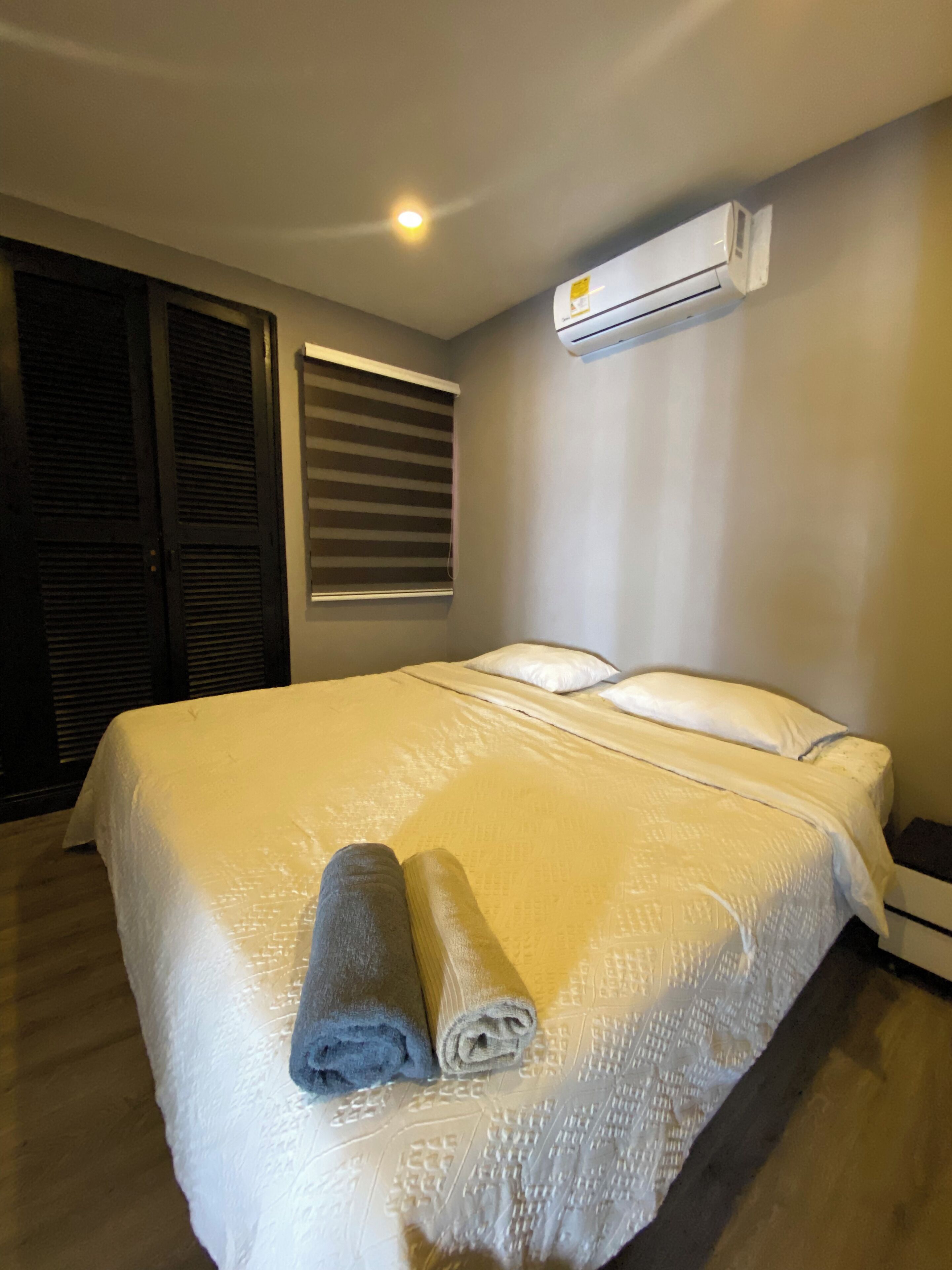Een net opgemaakt bed met een opgerolde handdoek, een raam met zonwering en een airconditioner aan de muur.