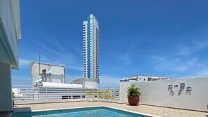 Une piscine sur le toit, avec un ciel bleu dégagé, un grand immeuble en arrière-plan et un mur blanc orné d’une plante en pot.