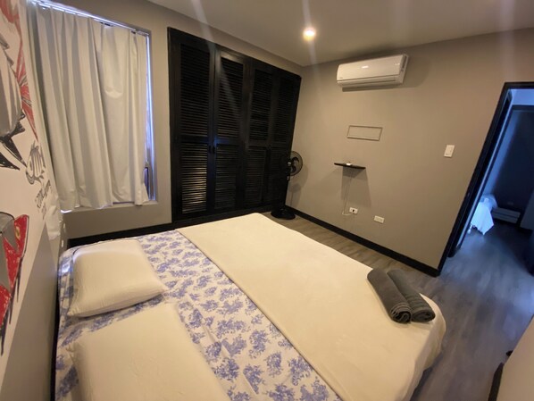 Une chambre d’hôtel avec un lit, une fenêtre avec des rideaux, un climatiseur fixé au mur et une porte donnant sur une autre pièce.