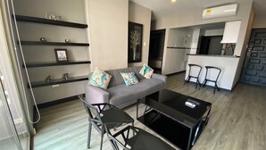 Un salon moderne avec un canapé gris, une table basse noire et des fauteuils assortis. On y trouve également un coin cuisine avec un réfrigérateur blanc et une table à manger avec des chaises. Des étagères ornées d'objets décoratifs sont visibles sur le mur de gauche.