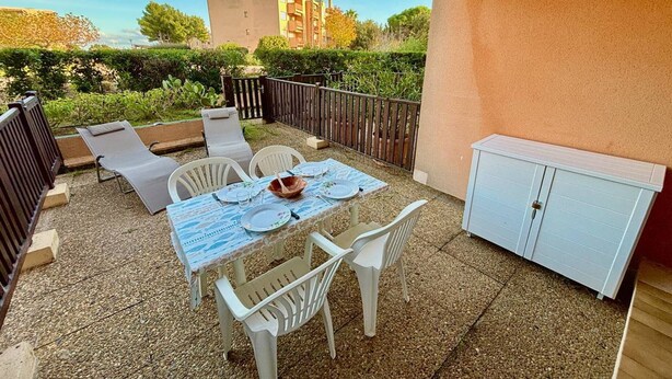 Une terrasse avec une table dressée pour le repas, des chaises blanches, un buffet blanc et, en arrière-plan, une vue sur un bâtiment.