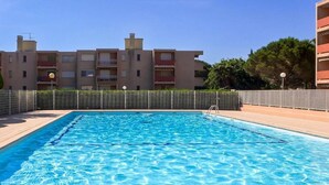 Une piscine rectangulaire à l’eau d’un bleu limpide, entourée d’une terrasse carrelée et d’une petite clôture. Au fond, on aperçoit des immeubles à plusieurs étages dotés de balcons, et un ciel dégagé.