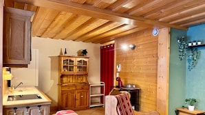 Um quarto aconchegante com paredes e teto de madeira, um armário de madeira com portas de vidro, uma cadeira vermelha, uma pequena mesa de madeira e uma cama com colcha estampada em vermelho e branco.