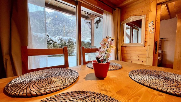 Interior de uma cabana de madeira, com mesa de jantar, cadeiras e vista para montanhas cobertas de neve através de grandes janelas.