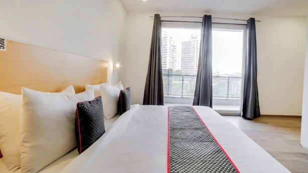 A hotel room with a large bed, a headboard, and a view of buildings through the window. - FLORA RESIDENCY (Gurugram)