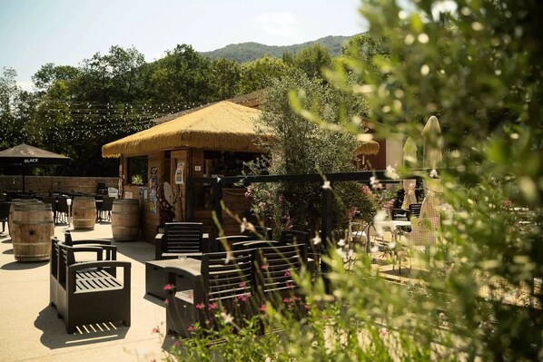 Un espace extérieur aménagé avec des tables, des chaises et des tonneaux en bois, entouré de verdure et bordé d’un bâtiment à toiture de chaume.