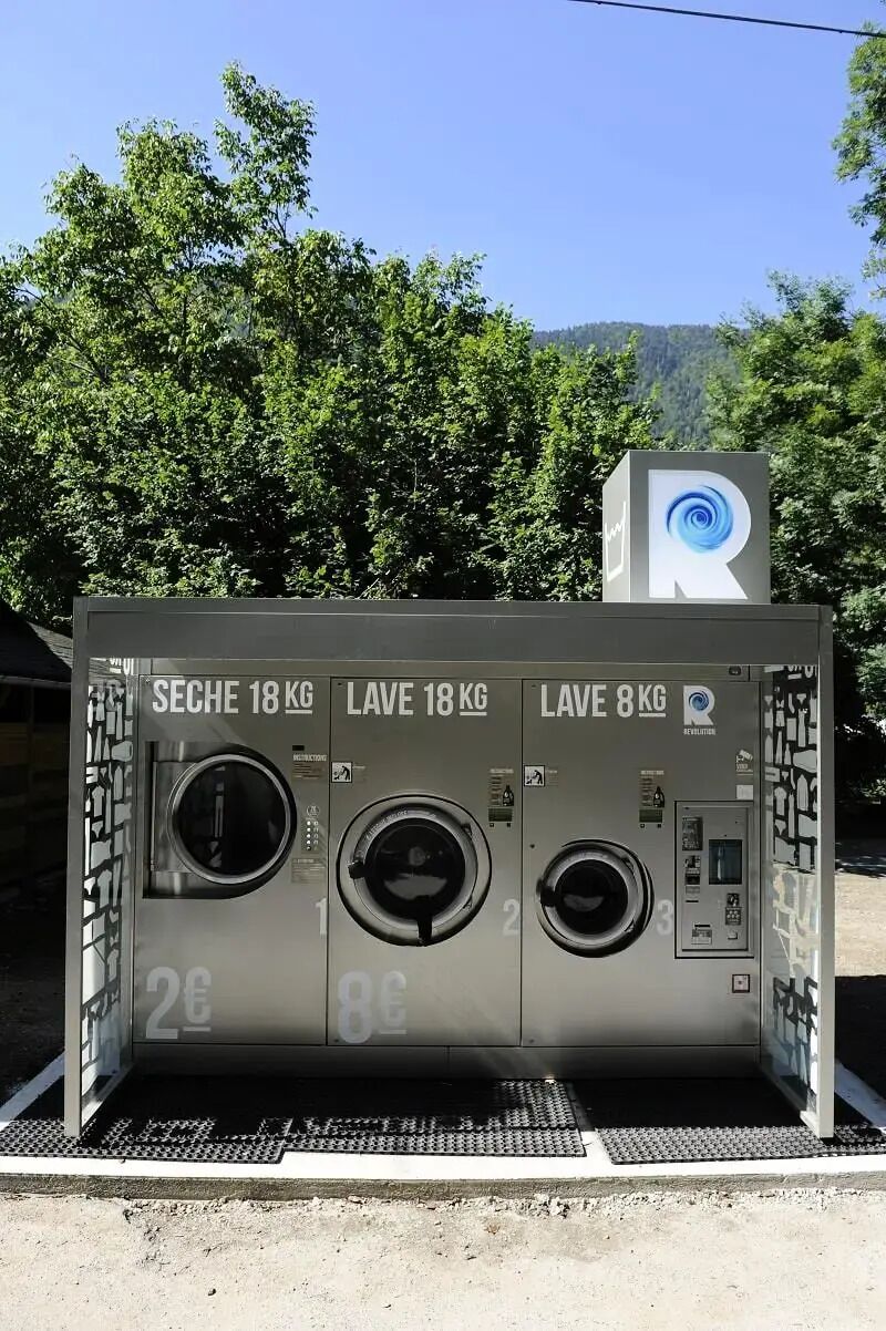 Une laverie publique équipée de trois machines à laver, dont trois sont étiquetées « SÉCHOIR 18 KG », « LAVE-LINGE 18 KG » et « LAVE-LINGE 8 KG ».