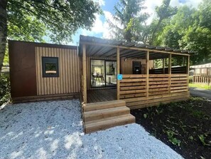 A wooden cabin with a covered porch and a small outdoor seating area. - Premium Mobile Home, 40 m² - 2 Bedrooms + Covered Patio + Air Conditioning + TV, Sleeps 4 (AX LES THERMES)