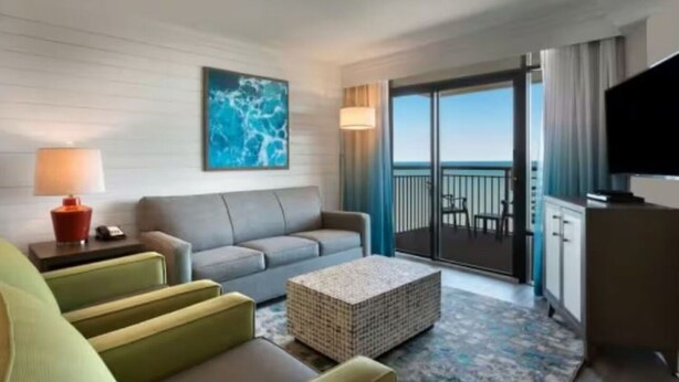 Habitación de hotel con sofá gris, una mesita de centro, televisor y balcón con vistas.