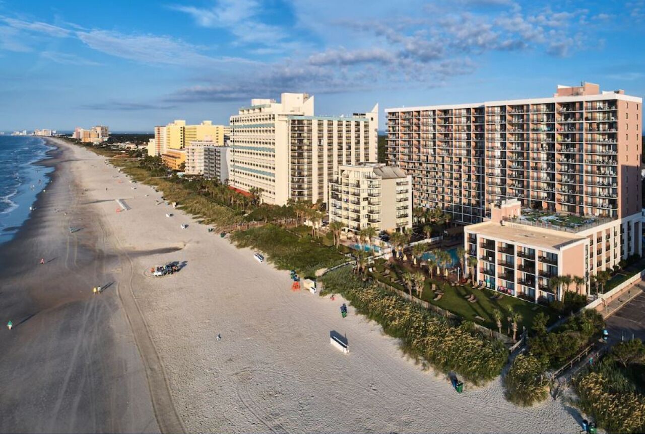 Un hotel frente a la playa con varios edificios altos, una playa de arena y cielo despejado.