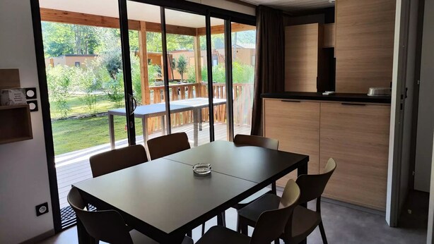 A modern kitchen with a wooden countertop and cabinets, a dining area with a table and chairs, and a sliding glass door leading to a deck with a view of greenery and buildings. - Chalet SPA Premium 42m² - 2 bedrooms + Covered terrace + Reversible air conditioning + TV 4 people (AX LES THERMES)