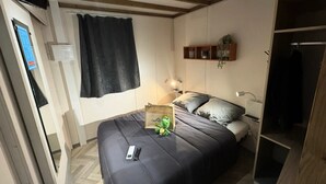 A bedroom with a bed, bedside table, lamp, and a small shelf with decorative items. - Chalet SPA Premium 42m² - 2 bedrooms + Covered terrace + Reversible air conditioning + TV 4 people (AX LES THERMES)