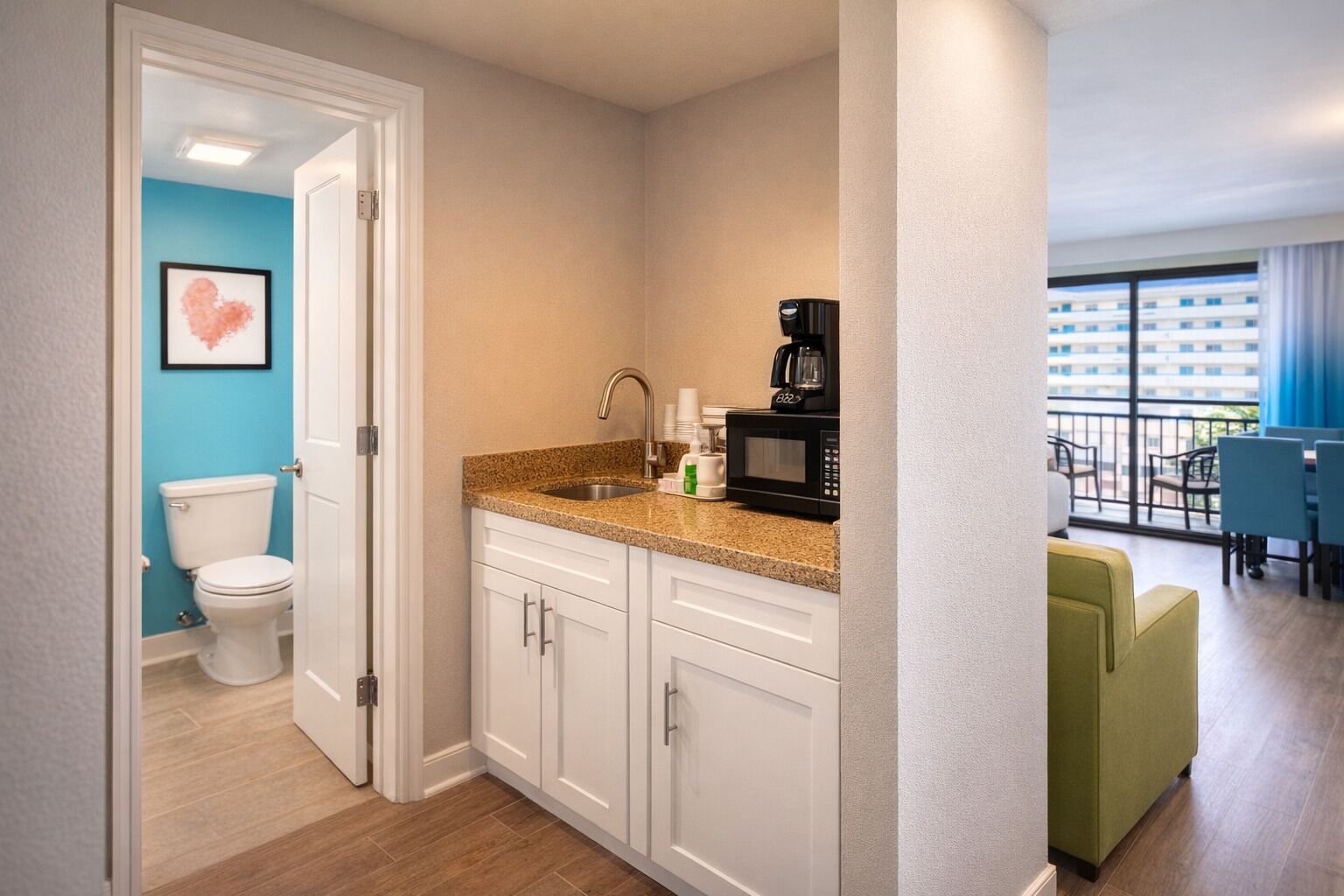 A compact kitchen with a sink, microwave, and coffee maker, adjacent to a bathroom with a toilet and a framed picture on the wall.