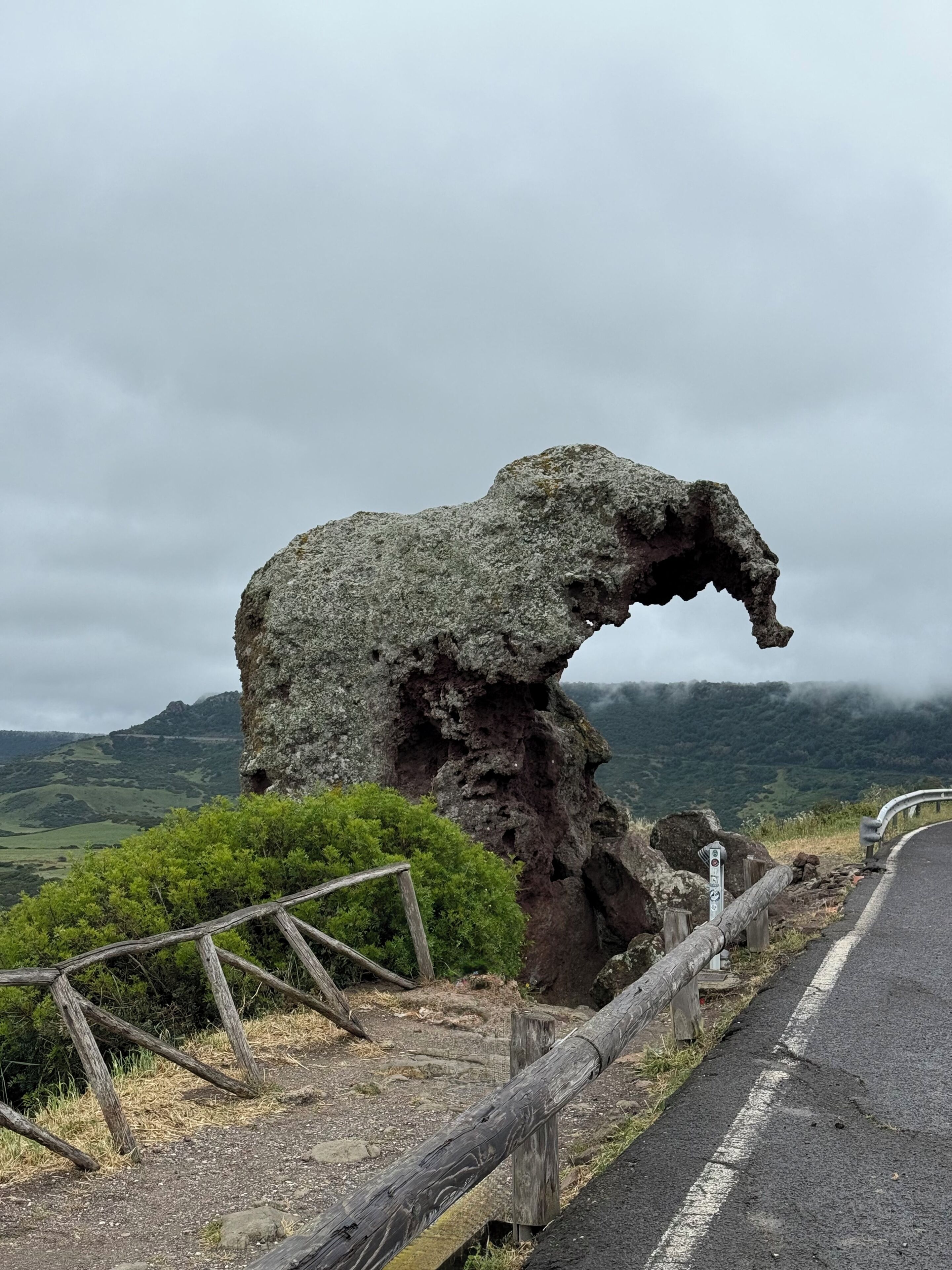 Eine natürliche Felsformation mit gewölbter Überhänge, eine gepflasterte Straße mit Schutzrail und eine gebirgige Landschaft im Hintergrund.