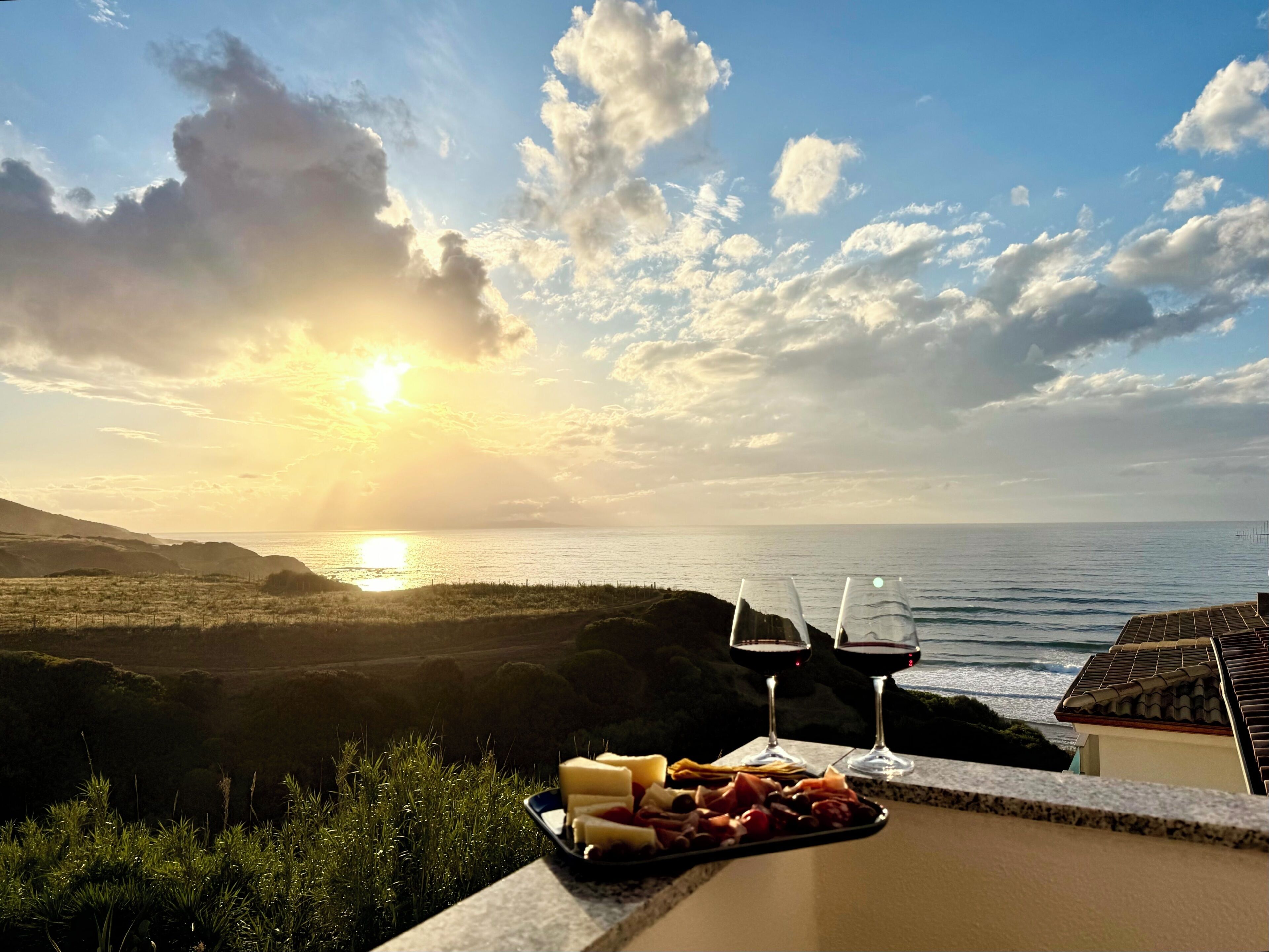 Een terras met uitzicht op zee en zonsondergang, een dienblad met eten en twee glazen rode wijn.