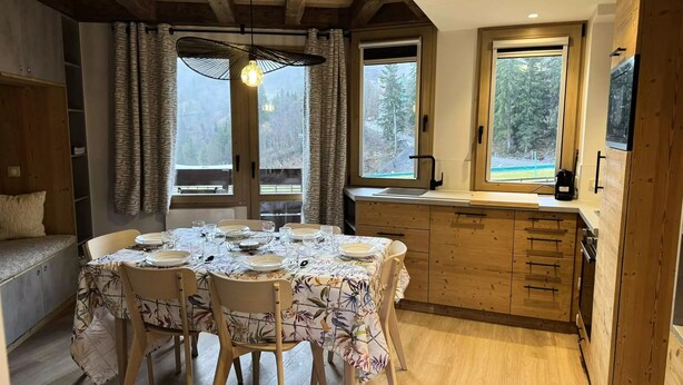 A dining area with a table set for four, wooden cabinets, and large windows with curtains. - Le Morel 35 - Lovely apartment completely renovated (LES AVANCHERS VALMOREL)