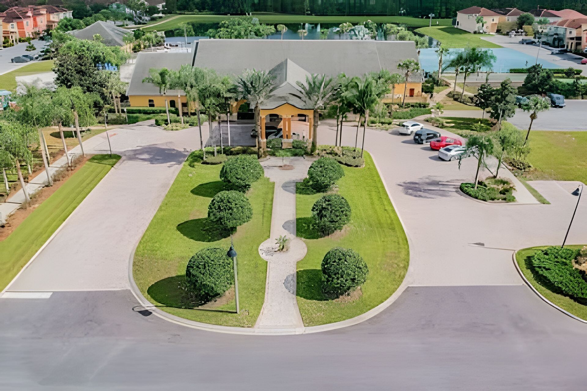 An aerial view of a gated community with a central building, surrounded by well-maintained lawns and a parking area with several cars.