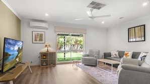 A modern living room with a grey sofa, a small armchair, a coffee table, a TV on a wooden stand, and a large window with a view of greenery. - Charming 3-bed Family Home With Amazing Garden (Melbourne)