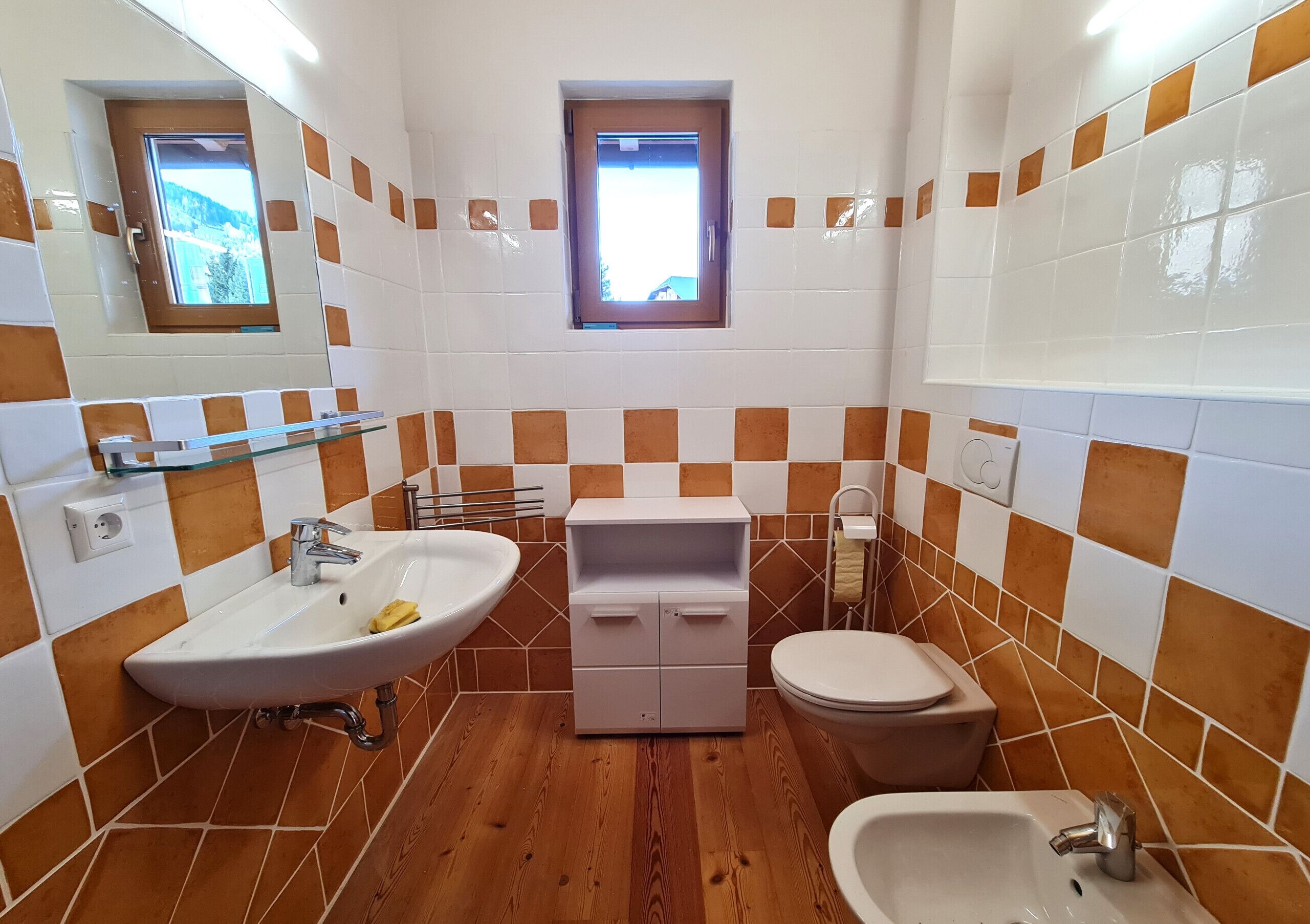 Ein Badezimmer mit Holzboden, weiß-orangenen Fliesen und einem Fenster. Es enthält ein Waschbecken, eine Toilette und einen kleinen Schrank.