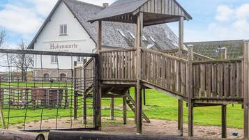 Una estructura de juego de madera con pared de escalada y zona cubierta, junto a un edificio blanco con el nombre 'Hohenzwarth' a la vista.