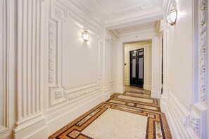 Un pasillo con paredes blancas ornamentadas, un suelo con diseño y un espejo que refleja la puerta al fondo.