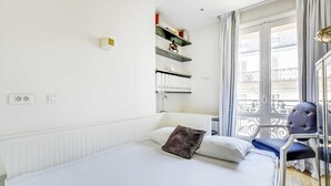 A hotel room with a bed, a chair, a window with a view of buildings, and a wall-mounted shelf with decorative items. - Bright Apartment - 2br/4p - Muette/passy - Trocadero (Paris)