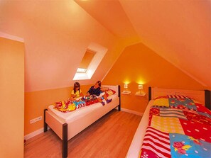 Ein Schlafzimmer mit schrägen Wänden, einem Bett mit farbenfroher Steudecke, einem Nachttisch mit Lampe und einem kleinen Fenster.