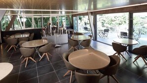 Een modern interieur met ronde tafels en stoelen, grote ramen met uitzicht op groen, en een houten plafond.