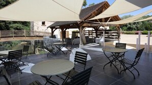 Een terras buiten met ronde tafels en stoelen, beschaduwd door grote parasols, en een stenen gebouw op de achtergrond.