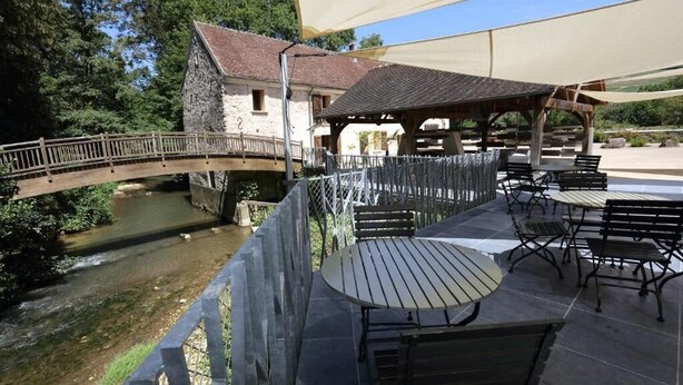 Een terras aan de rivier met buitenmeubilair, een stenen gebouw en een houten brug.