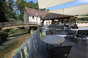 A riverside patio with outdoor seating, a stone building, and a wooden bridge. - Le Moulin Babet (Mezy-Moulins)