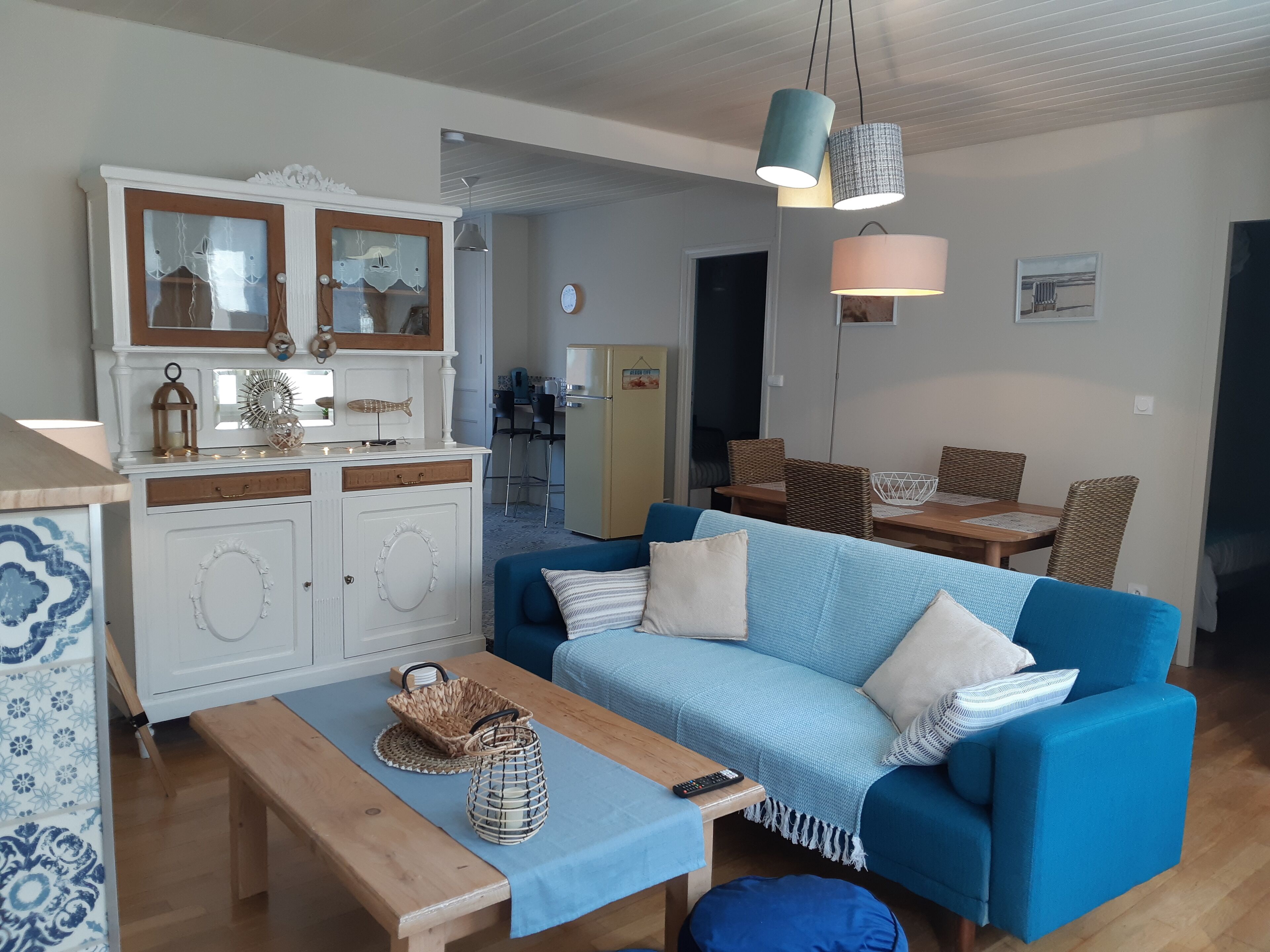 A cozy living room with a blue sofa, wooden coffee table, and a kitchen area with a white cabinet and a refrigerator.