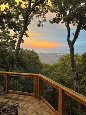 Une terrasse en bois avec une rambarde en métal, donnant sur un paysage boisé au coucher du soleil.