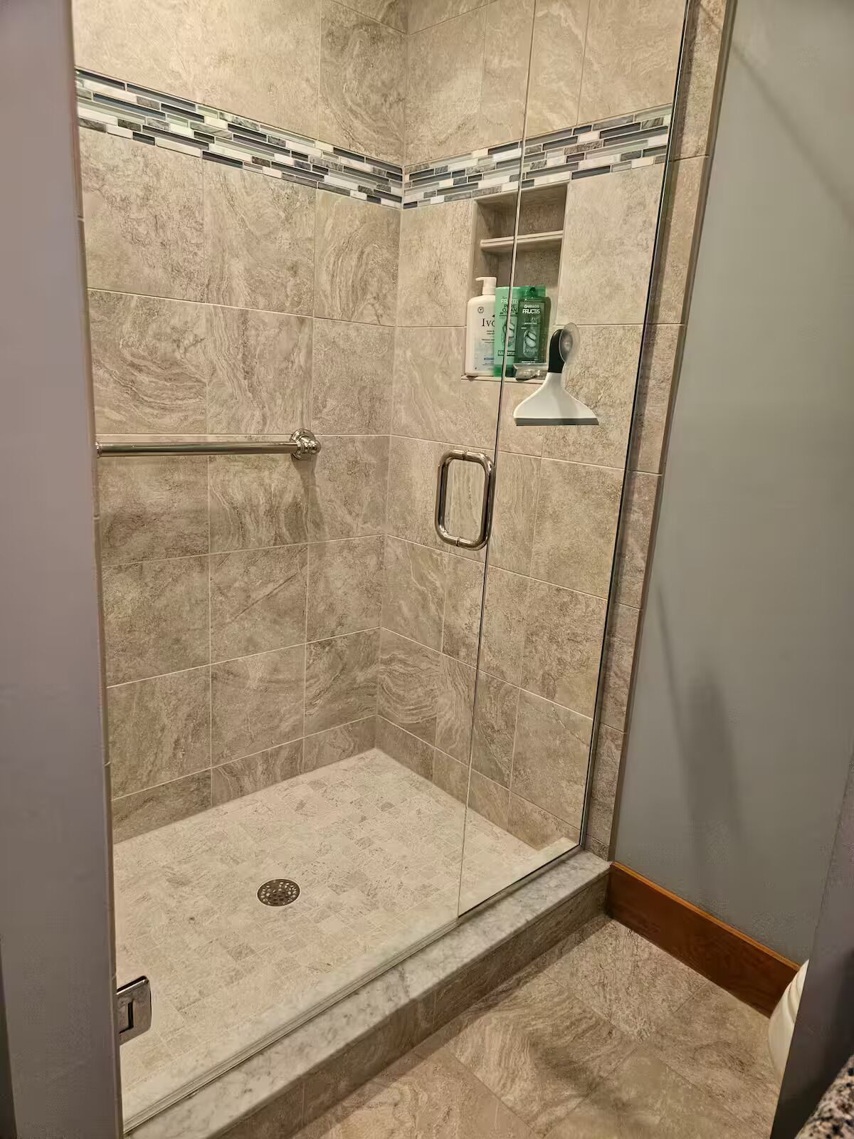 A glass-enclosed shower with beige tiles, a metal handrail, and a small shelf with toiletries.