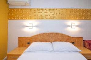 Habitación de hotel con cabecera de madera, dos lámparas de noche, una cama con ropa blanca y un equipo de aire acondicionado sobre la cama.