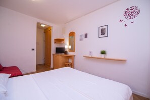 Ein Schlafzimmer mit einem Bett, einem Schreibtisch, einem Fernseher, einem Spiegel und einem Regal mit einer Pflanze.